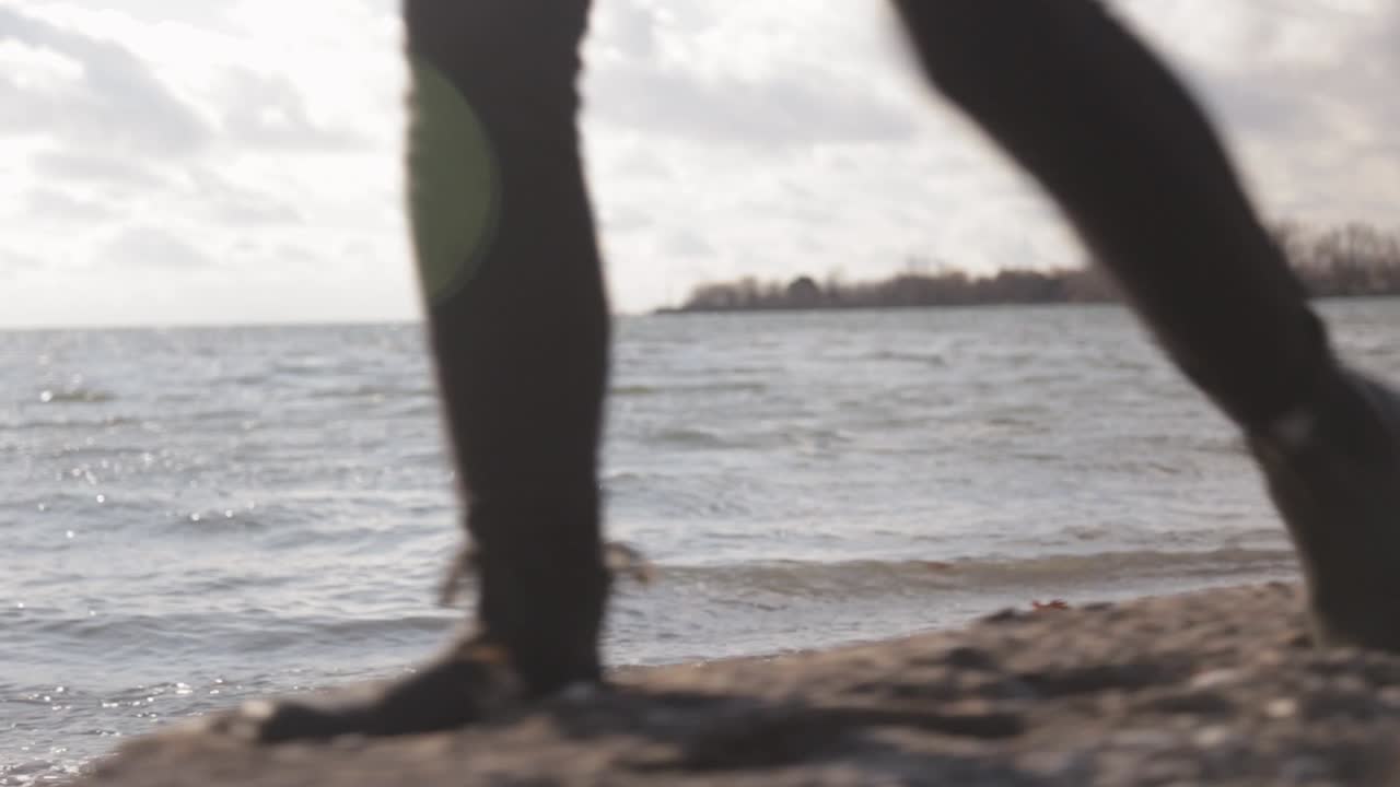 Back Side View Of A Woman Feet Walking Along The Lake Shore With Waves Crashing To The Shore During Sunny Day - Close Up Shot