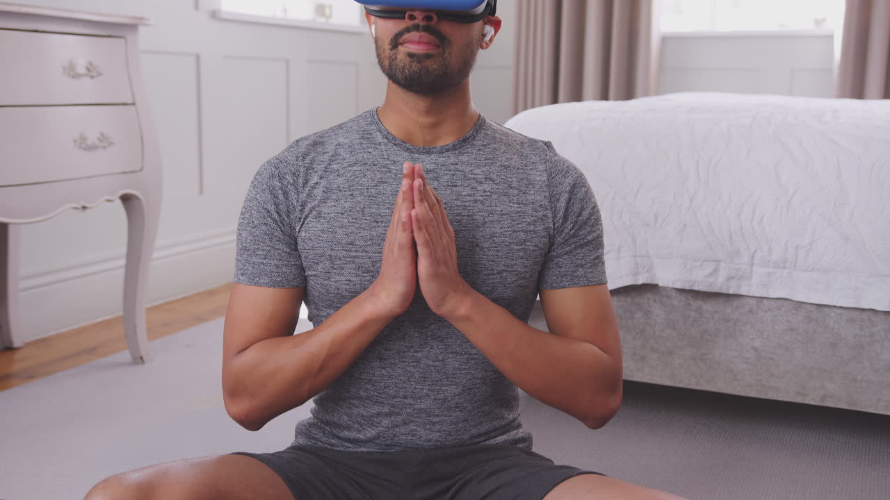 Man sitting on yoga mat in bedroom at home wearing virtual reality headset and wireless earphones and streaming virtual class from mobile phone - shot in slow motion