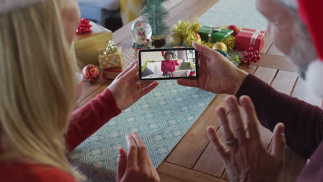 pareja caucásica con sombreros de santa usando un teléfono inteligente para una videollamada de navidad con una mujer en la pantalla