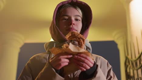 young woman standing outdoors with glowing streetlight in background, holding crispy pastry in hand, warm city light illuminates her face