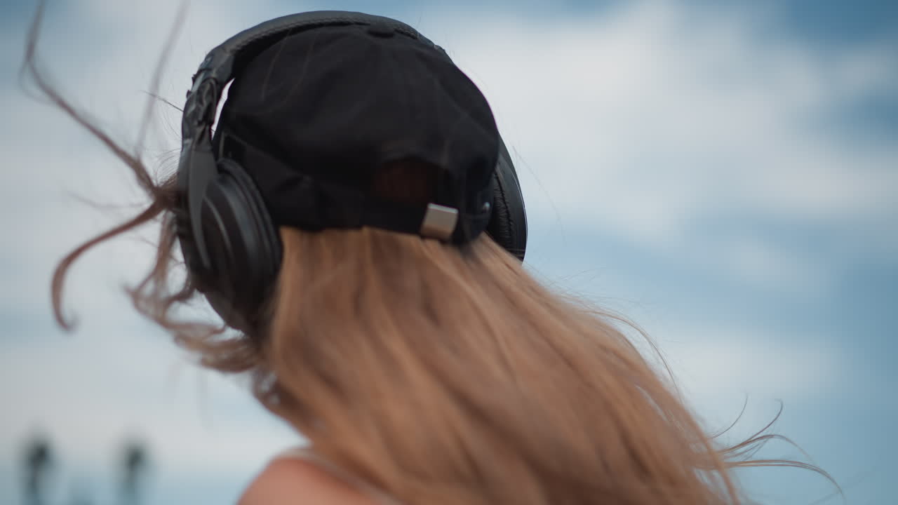 Giovane donna bianca con le cuffie in strada, che sorride e balla, si volta verso il cielo, si aggiusta il cappello e le cuffie, capelli che si muovono nella brezza, atmosfera estiva, ritratto spontaneo che trasmette gioia