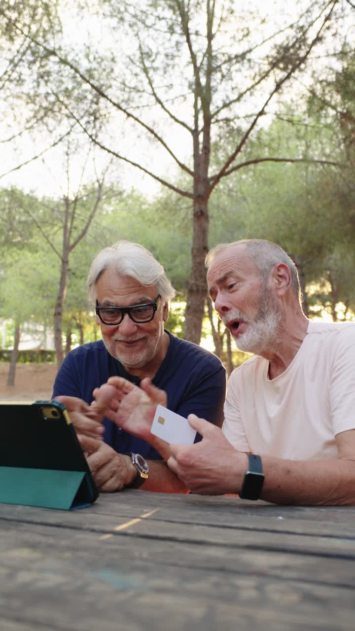 Due uomini anziani che usano un tablet e una carta di credito in un parco