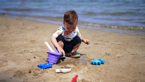 adorable toddler boy plays with toys at the seashore. kid pours some water from his bucket on the sand.