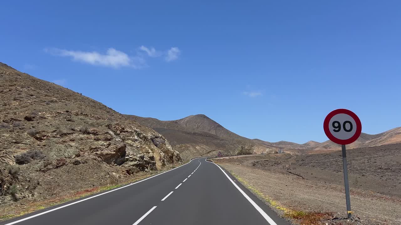 POV driving through the scenic volcanic mountains near Betancuria, Fuerteventura, passing speed limit sign on a sunny day. Canary Islands, Spain