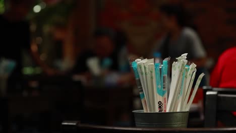 a collection of straws in a container with a blurred restaurant background, captured in natural lighting