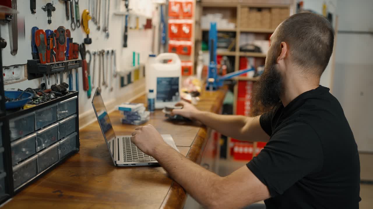 maestro caucásico usando una computadora portátil en un taller de bicicletas, eligiendo parte de la bicicleta en línea