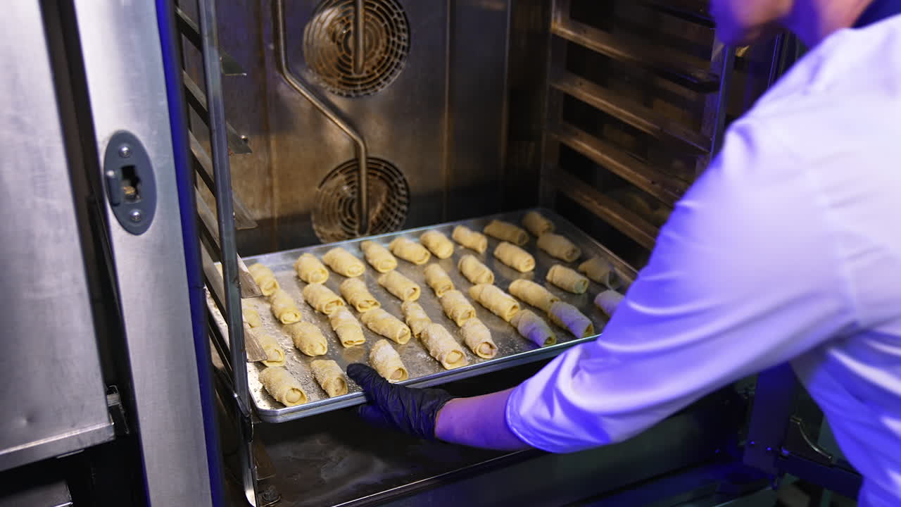 Baker puts three baking sheets filled with little croissants into the oven. Sweet pastry production at modern bakery.