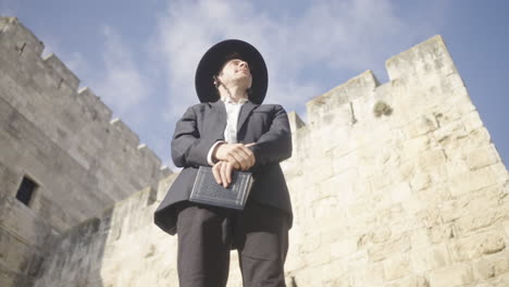 jerusalem old city, israel, an orthodox jewish man stands in front of the city walls wearing a black suit and hat and holding a prayer book on a sunny day