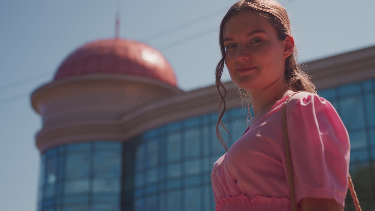 Mujer joven con vestido rosa paseando. La secuencia muestra a una estudiante paseando, una soñadora protegiéndose los ojos, una modelo posando con seguridad, una sonrisa en primer plano. Bolso de mimbre, pelo trenzado, barandilla de hierro forjado, edificio con cúpula de cristal.