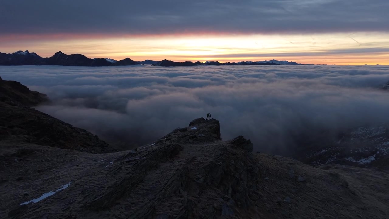 Sorvolo aereo su escursionisti che si godono il bagliore dell'alba sopra l'inversione delle nuvole nelle Alpi svizzere, con una panoramica verso l'alto delle montagne e del cielo colorato all'alba.