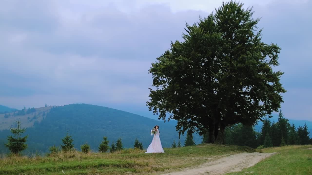 Woman in wedding dress on nature background. Young bride in white dress and hat standing near the green tree in the mountains.
