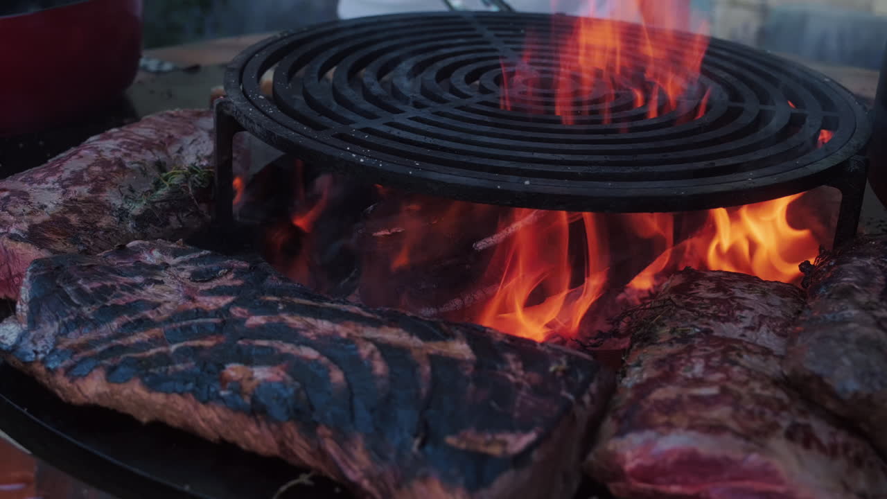 Big pieces of meat grilling on a barbecue with hot flames during the day, medium shot