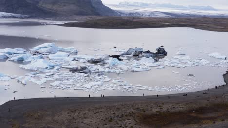 tourist people standing on glacial lagoon shore looking at calved icebergs, aerial