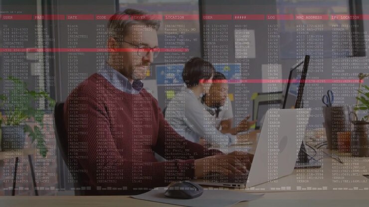 Mand i forgrunden taster på bærbar computer, igangsætter kodeoverlejring, der fylder rammen, team konsulterer telefon for IT