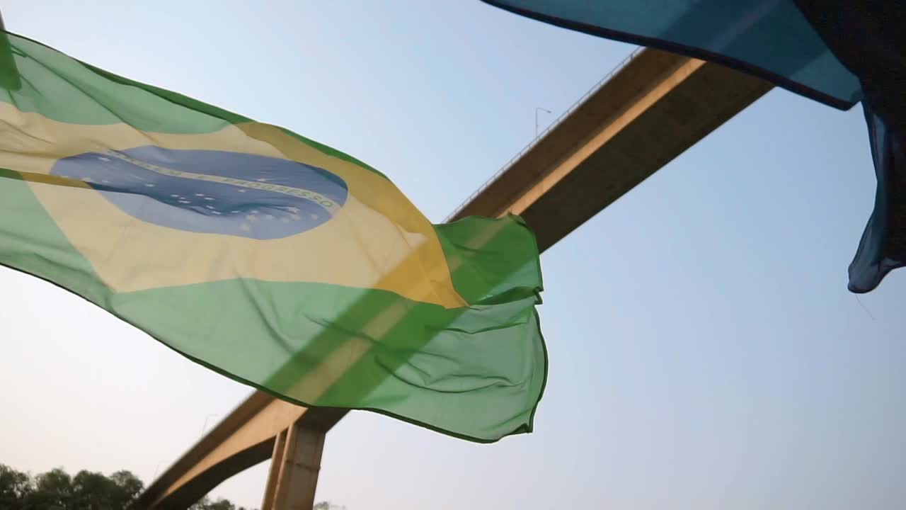las banderas nacionales de brasil y paraguay ondean en el viento, bailando armoniosamente contra el telón de fondo del puente que conecta brasil y argentina