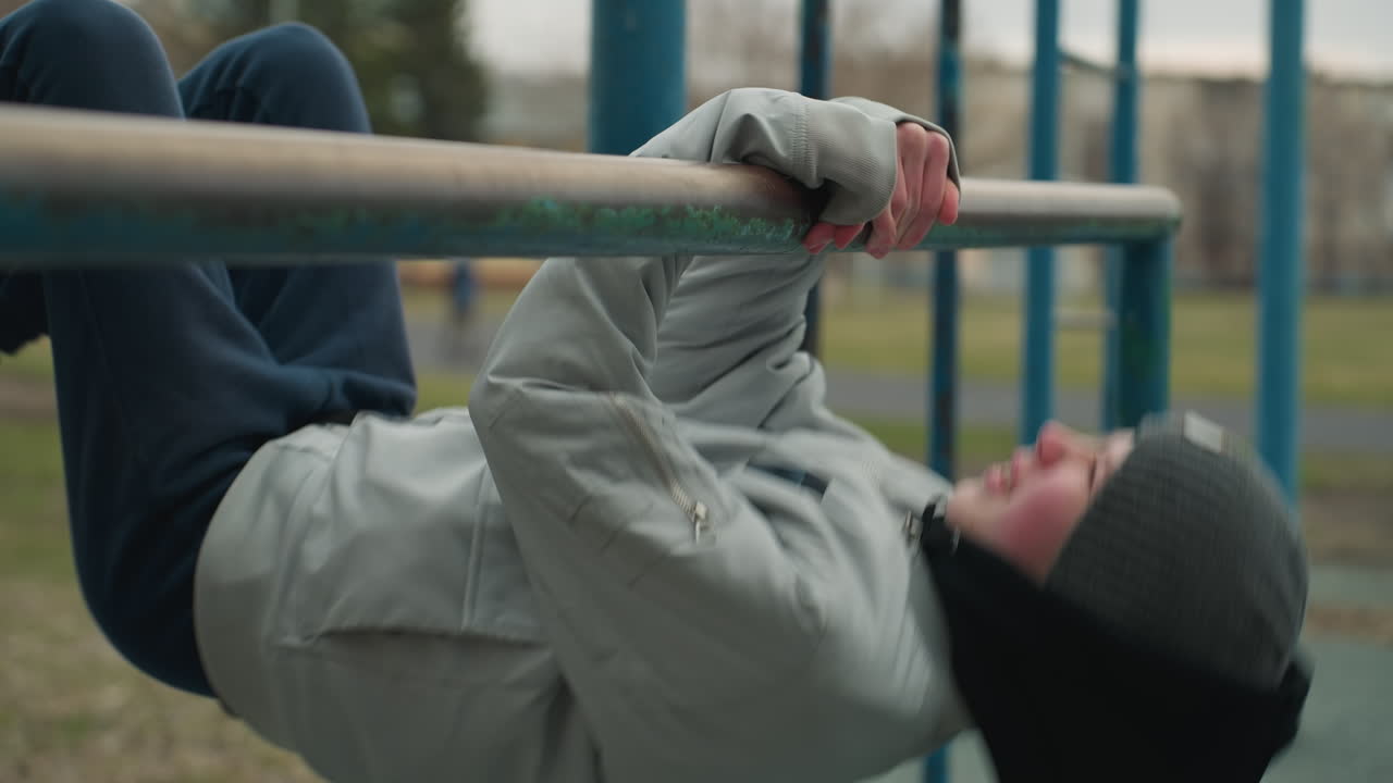 Close-up of a young boy exercising on an iron bar in the stadium, he is wearing a gray jacket and beanie