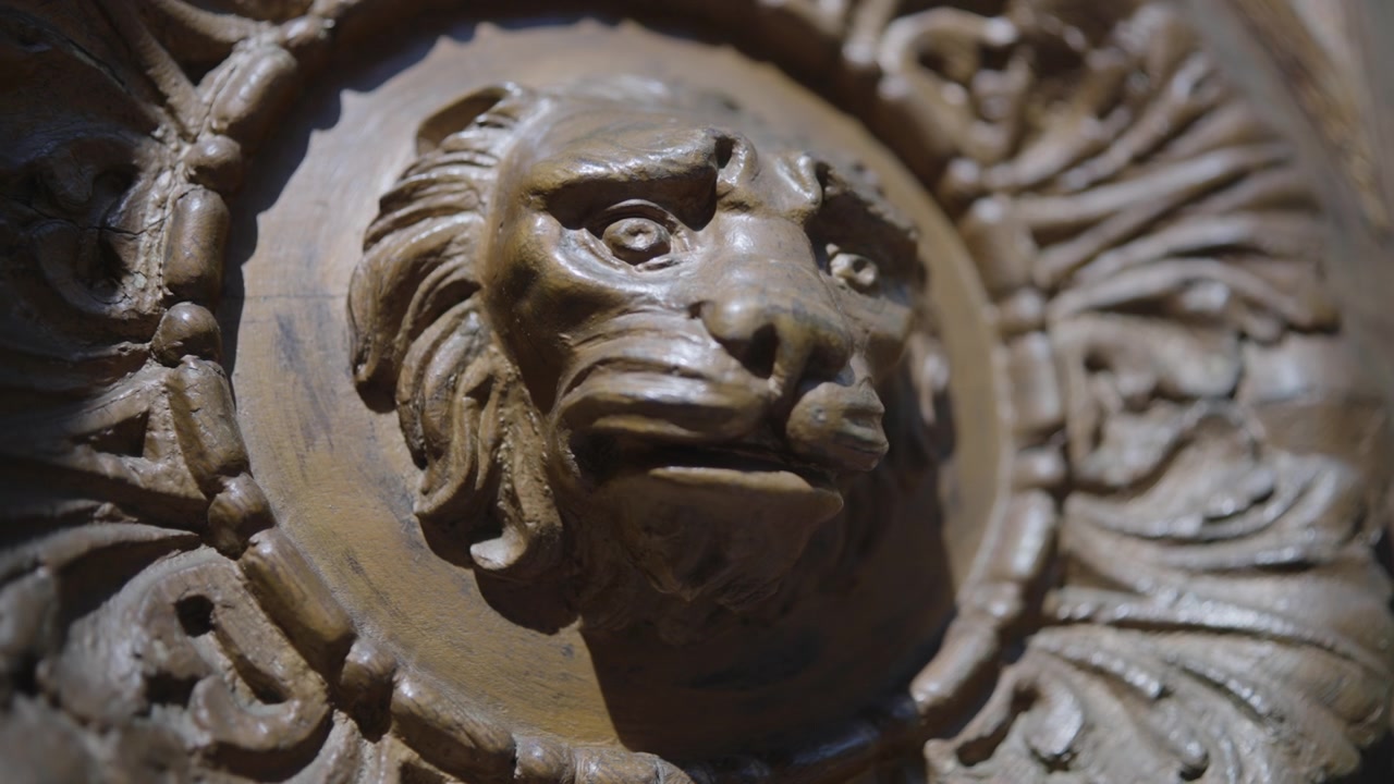 Vista ravvicinata orizzontale e dettagliata di un medaglione di leone in legno intagliato su un pannello decorativo, con un leggero movimento del punto di vista che rivela venature ricche, segni degli attrezzi, una calda patina, intarsi ornamentali e profondità.