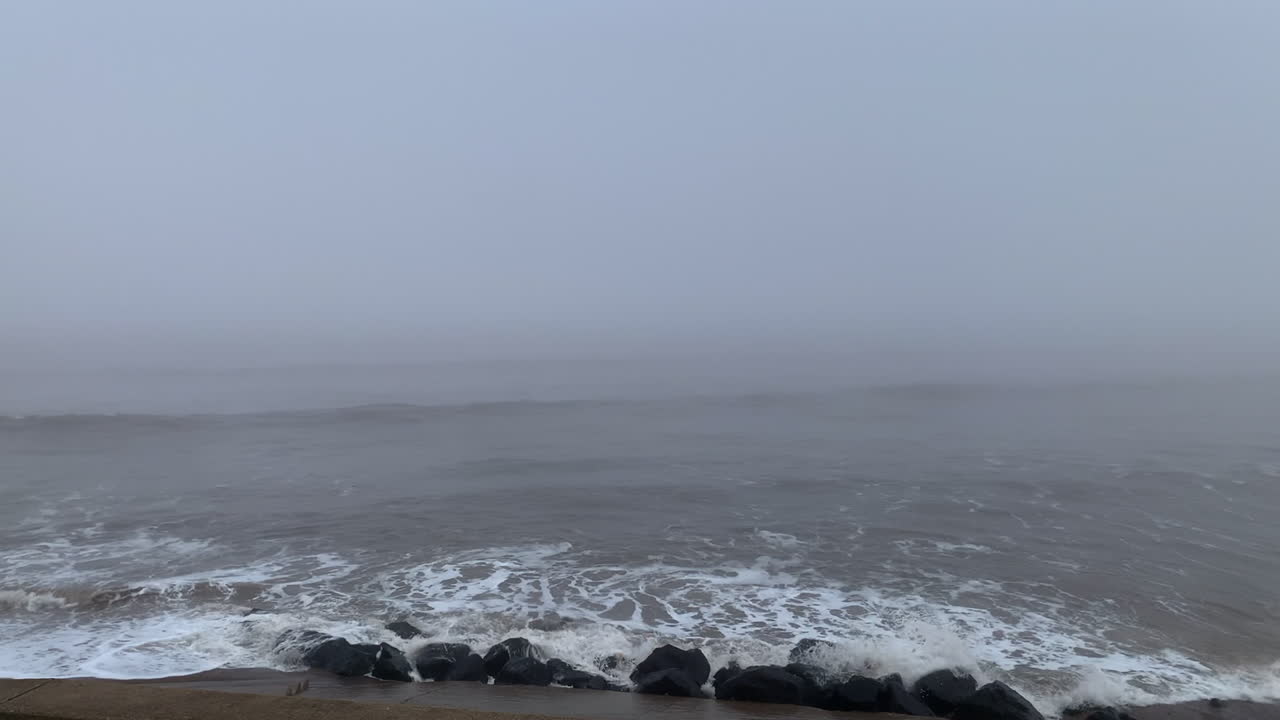 Mist rolling in from the ocean as waves crash against rocks under a grey foggy sky and stormy sea
