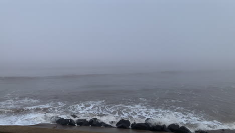 mist rolling in from the ocean as waves crash against rocks under a grey foggy sky and stormy sea