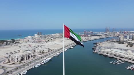 4k: drone camera view of the flag of united arab emirates waving in the air, the blue sky and city development in background, the national symbol of uae over sharjah's flag island