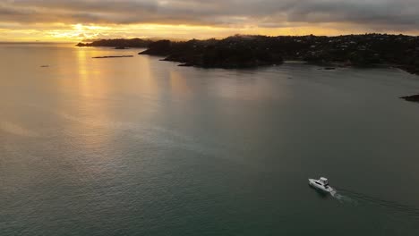 aerial establishing shot of speedboat cruising on tranquil waters along coastline of waiheke island, new zealand. golden sunset with clouds at sky. wide shot.
