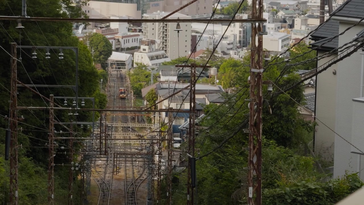 Wide shot perspective of steep funicular railway tracks cutting through lush greenery and residential houses, leading down toward the urban cityscape and buildings of Ikoma.
