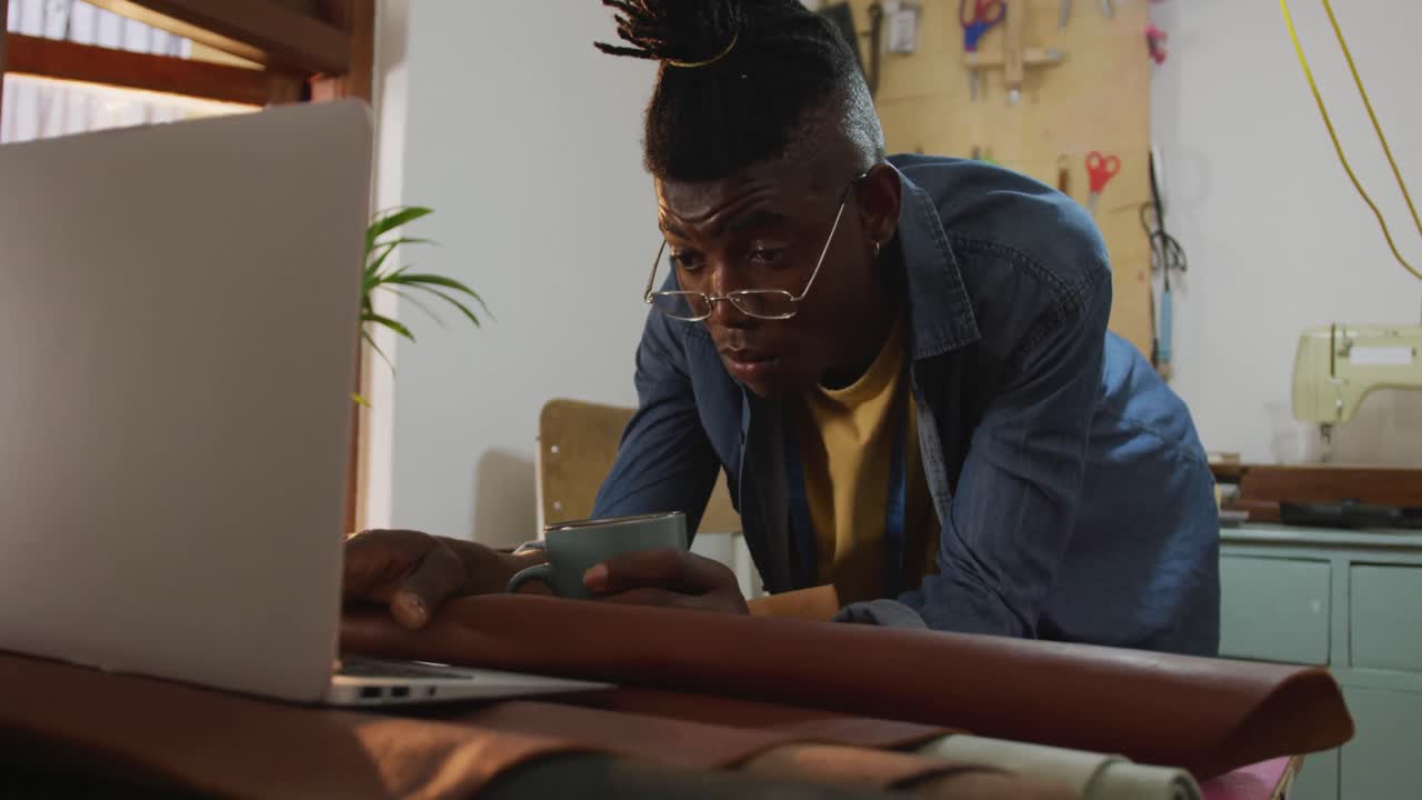 African american craftsman wearing glasses drinking coffee and using laptop in leather workshop