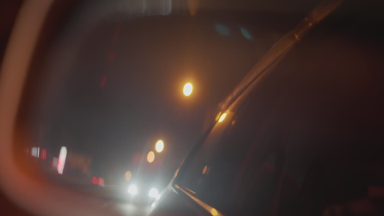 밤에 백미러에 다가오는 헤드라이트가 보이고, 택시 운전사는 도시 불빛이 흐릿해지는 동안 거울을 흘끗 봅니다. 호박색 가로등이 눈부심과 보케를 만들고, 긴장된 분위기 속에서 조심스럽게 운전하는 모습이 담겨 있습니다.