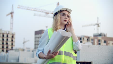 Woman-engineer-designer-talking-on-the-phone-with-the-contractor-with-drawings-in-hand-on-the-background-of-buildings-under-construction-and-cranes