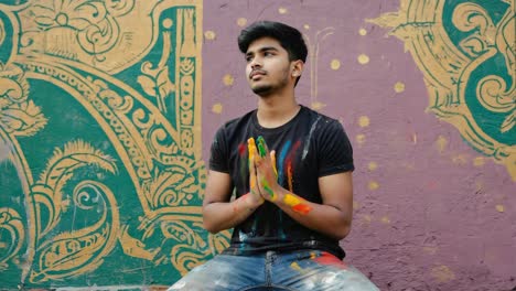 young artist with colorful hands joined in prayer looking up in front of a large mural, expressing gratitude, hope, or seeking inspiration after completing his artwork