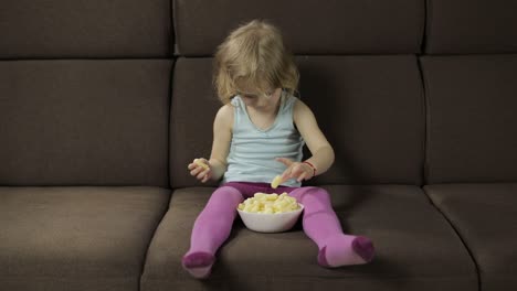 girl sitting on sofa and eating corn puffs. child smiling and taste puffcorns