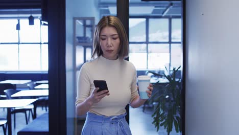 mujer de negocios asiática caminando usando un teléfono inteligente sosteniendo una taza de café en una oficina moderna