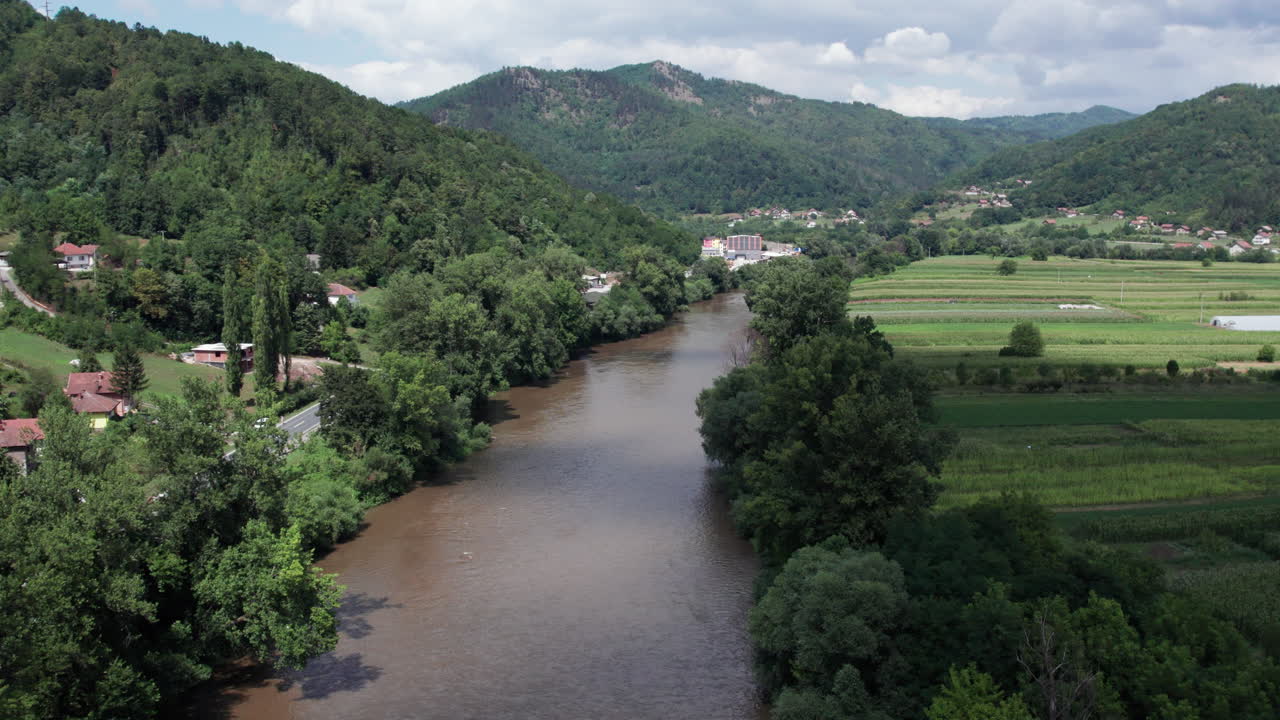Drone view of brown muddy river winding through farmland fields and forested hills