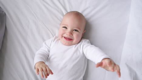 a child in a white t-shirt lying on a white bed looking at the camera and laughing in slow motion, top view