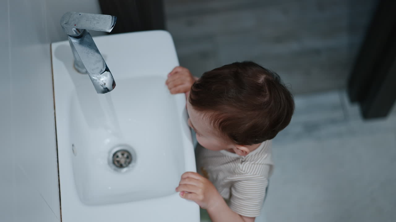 Caucasian toddler standing at the sink playing with faucet. Excited kid touches the water. High angle view.