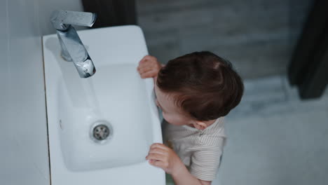 caucasian toddler standing at the sink playing with faucet. excited kid touches the water. high angle view.