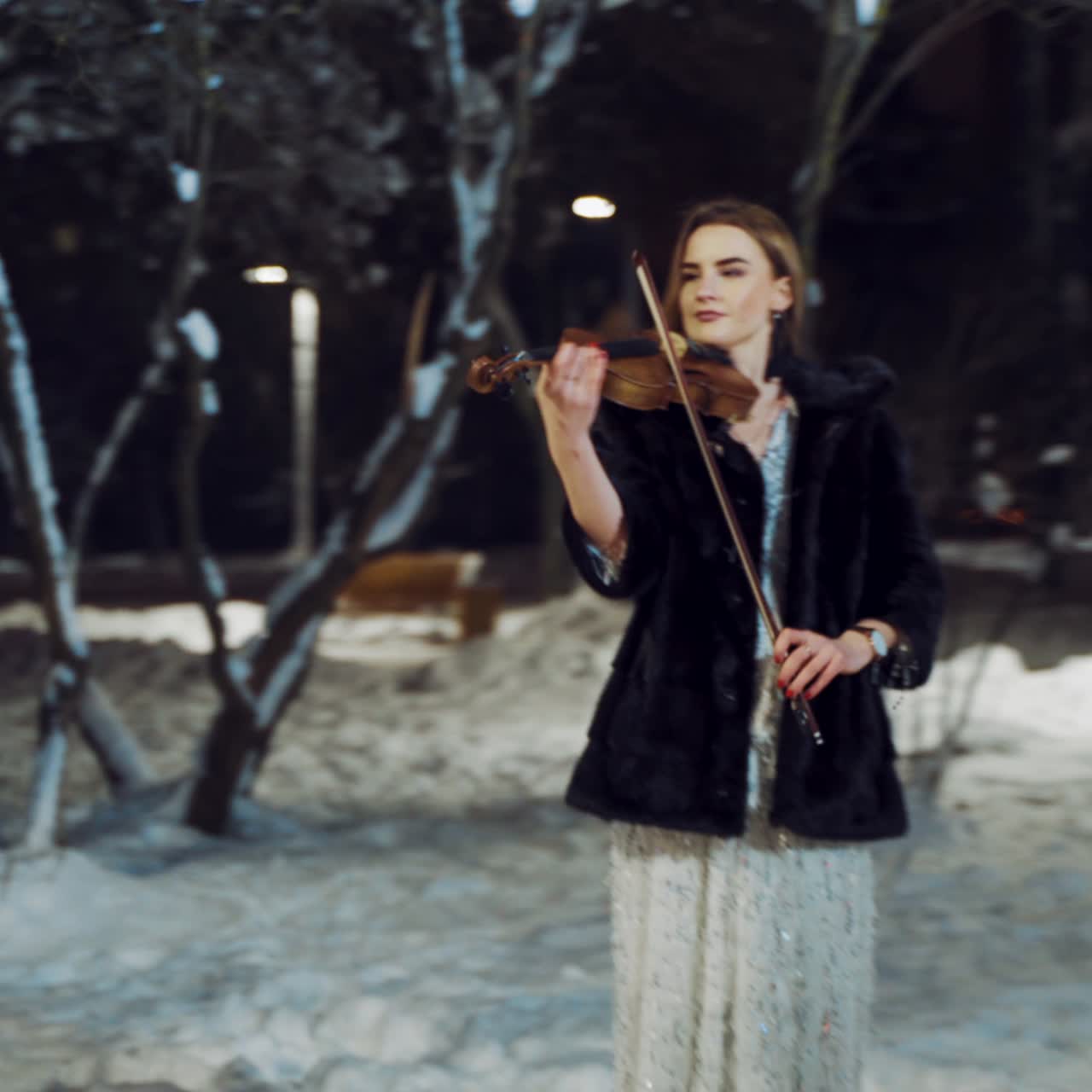 A gorgeous girl in a white dress with sparkles and in a fur coat is standing in the park and playing the violin in the winter on the background of trees in the evening. Camera motion to right