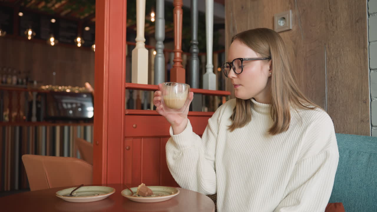 Side view of student in white sweater and glasses sipping latte from glass cup while seated at round table in cozy coffee shop with soft lighting and dessert plate beside empty saucer