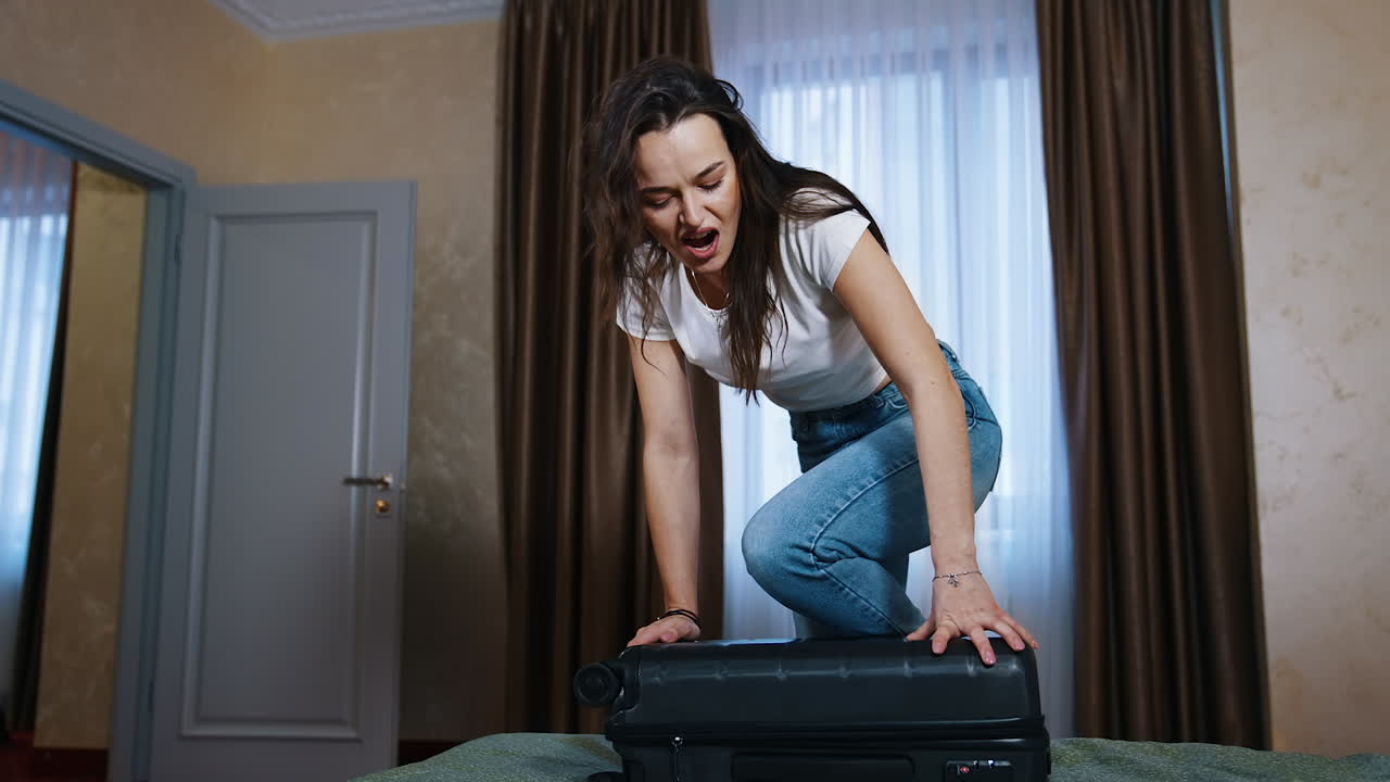 Young woman struggling to close full suitcase. Tired woman trying to close her travel luggage on bed in the hotel room.