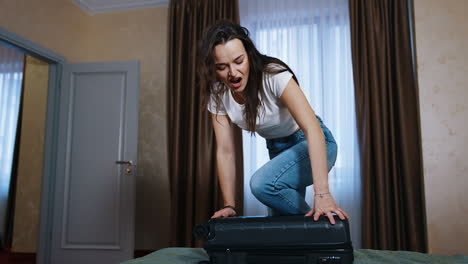 young woman struggling to close full suitcase. tired woman trying to close her travel luggage on bed in the hotel room.