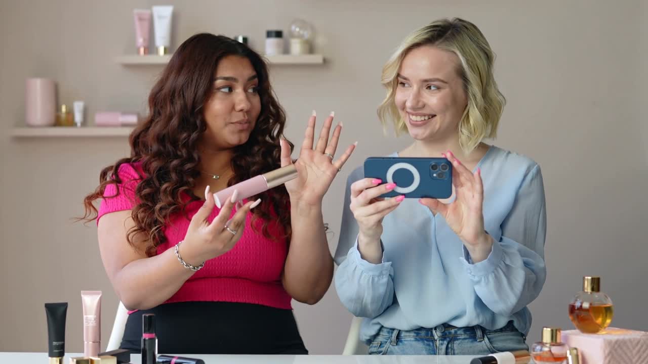 Angling blue phone ring, cohost filming host in pink top turning product at vanity, demoing
