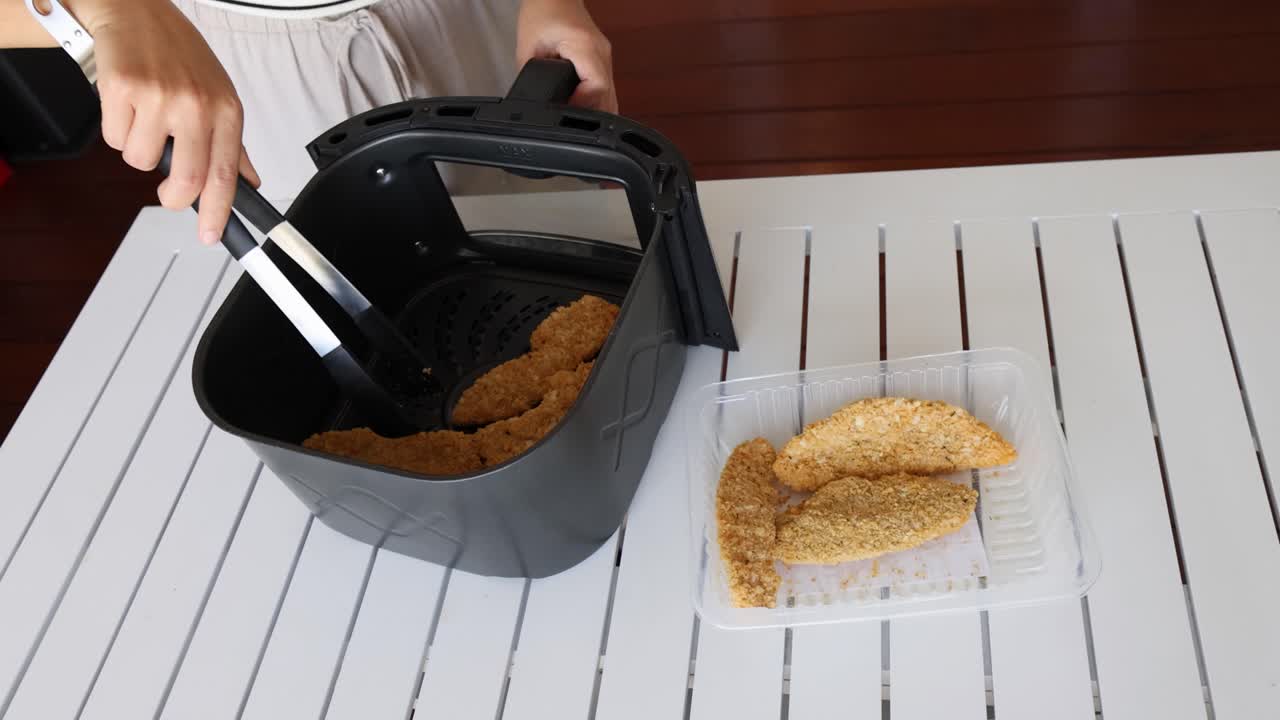 A person uses tongs to transfer cooked chicken from an air fryer to a tray on a white table