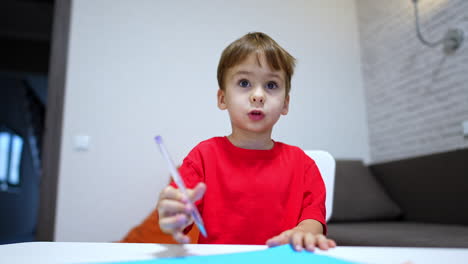 toddler kid in red t-shirt sits at desk holding a pen. cute baby boy talks to camera. low angle view.