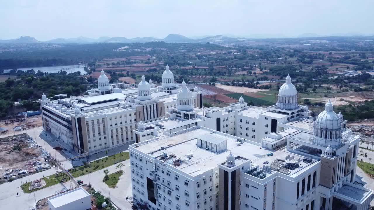 Aerial tracking shot over a massive, modern university or corporate campus with multiple white domes, set in a developing rural landscape with distant hills and a reservoir