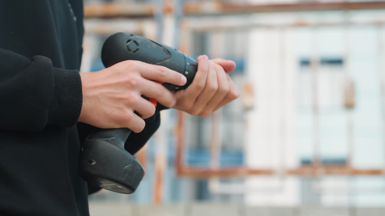 Closeup hands holding cordless drill prepping bit against scaffolded building exterior, focused grip and finger on trigger, battery indicator visible, urban renovation atmosphere, handson builder
