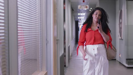 Smiling-biracial-businesswoman-talking-on-smartphone-walking-down-corridor-in-modern-office