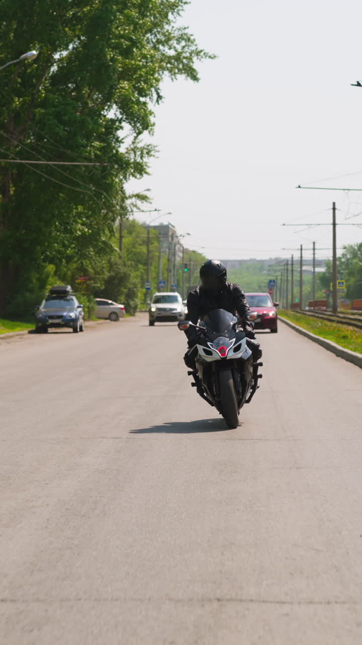 Skilled biker in black leather jacket rides fast motorcycle along street road with cars past tram route in modern city on sunny day slow motion
