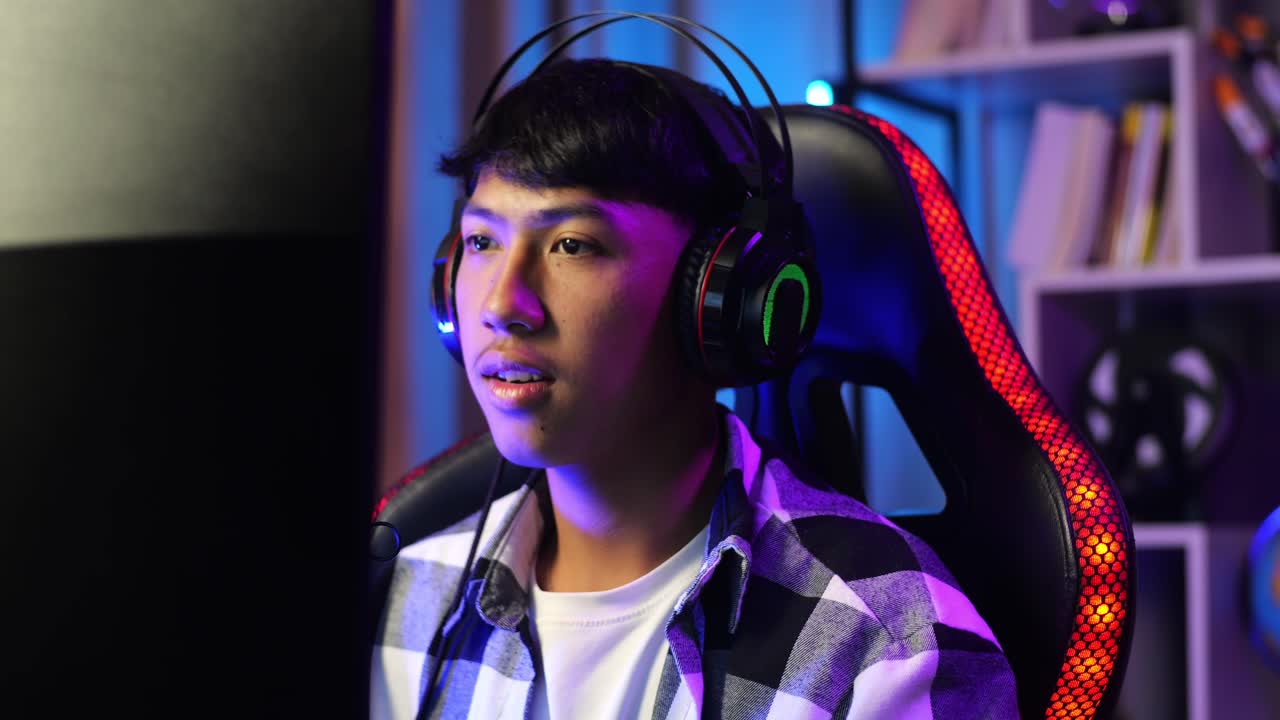 Primo piano di un ragazzo streamer asiatico che esulta e balla per celebrare la vittoria di una partita online sul suo computer. Videogioco in live streaming, con la scrivania illuminata da una striscia LED RGB.