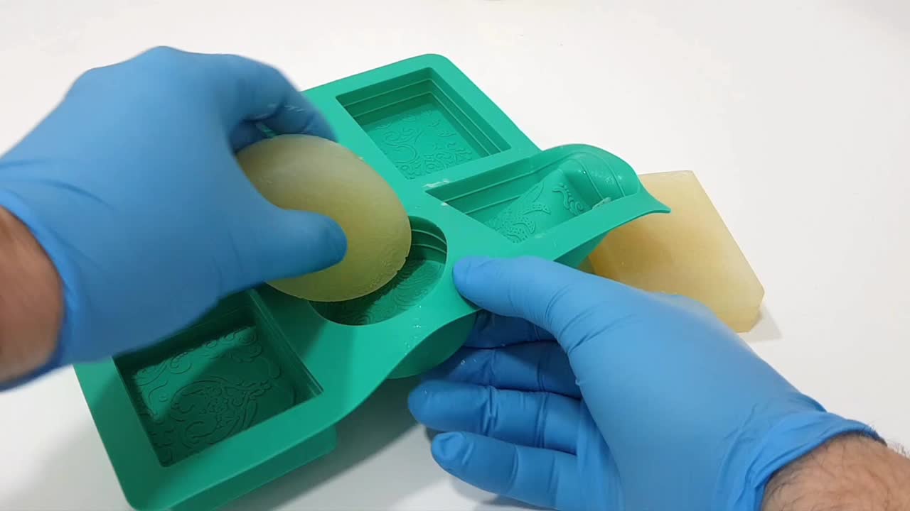 Close-up view of hands wearing blue gloves demolding freshly made soap bars using a silicone mold in a natural cosmetics laboratory.