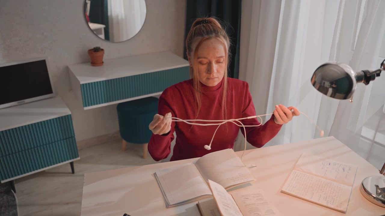 Jovem mulher de blusa vermelha está sentada à escrivaninha escrevendo. Ela desembaraça fones de ouvido brancos com fio enquanto manuseia vários itens. O cenário apresenta paredes de cores claras, carpete estampado e listras azuis.